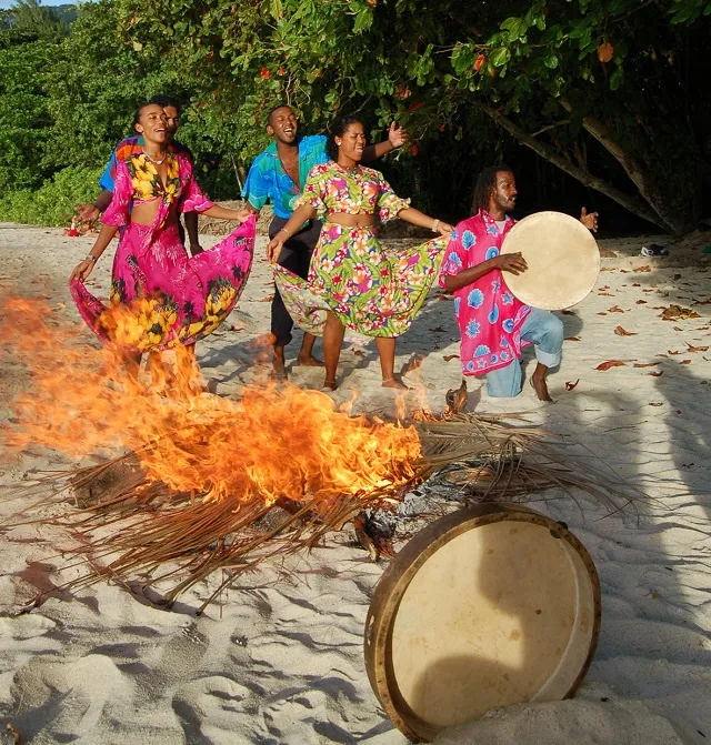 Tambour de moutya posé près d'un feu de palmes sur une plage des Seychelles, danseurs en arrière-plan
